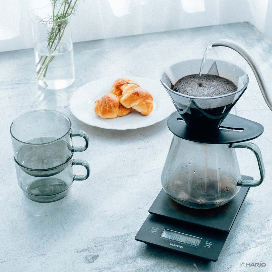 Coffee brewing process on a kitchen counter with Hario coffee maker, cups, and pastries.