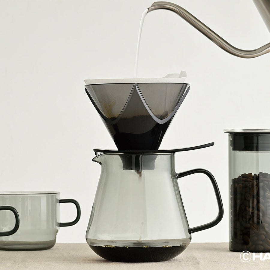 Coffee brewing process with a clear glass carafe and black coffee filter on a light background.