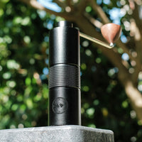 Black Hario Kabuto manual coffee grinder with wooden handle, displayed on a stone surface with natural greenery in the background – perfect for outdoor coffee brewing enthusiasts