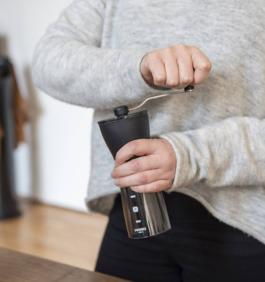 Person using the Hario Mini Mill PLUS manual coffee grinder, showing black top, crank handle, and transparent base with measurement markings.