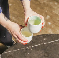 Two hands holding white Tajimi cups with green liquid on a stone surface.